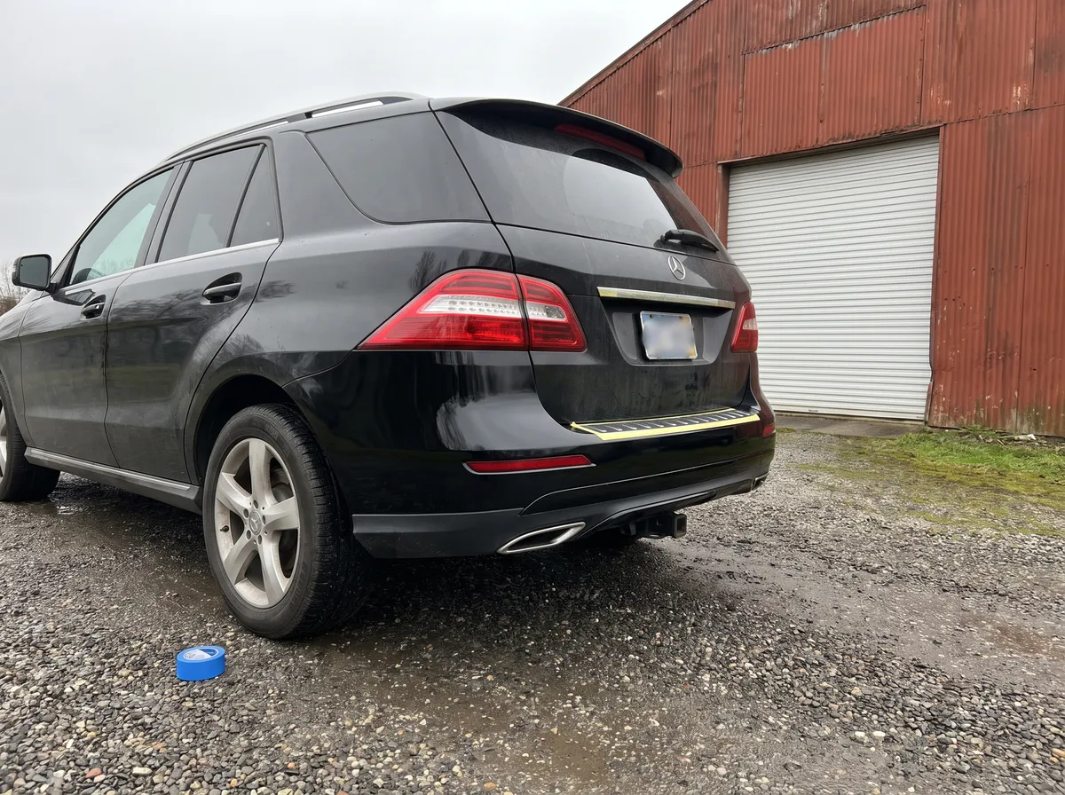 Svart Mercedes-SUV på grusplanen utanför en verkstad, med nylackerad bakre stötfångare och en rulle maskeringstejp kvar på marken